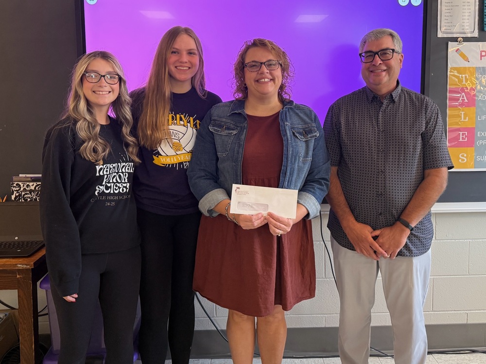 A photograph shows four people standing side-by-side in front of a classroom whiteboard displaying a solid purple light. On the left are two high school girls, smiling. The girl on the far left has blonde hair, glasses, and is wearing a black sweatshirt with "National Honor Society" text. The second girl has long brown hair and is wearing a dark t-shirt with a volleyball graphic. To their right are two adults. A woman in the center has curly brown hair, glasses, and is wearing a rust-colored dress with a denim jacket. She is holding a white envelope with two hands. On the far right is a man with gray hair, glasses, and is wearing a dark patterned short-sleeved shirt and light pants. The group appears to be in a classroom, with a desk and various posters or boards visible on the edges of the frame.