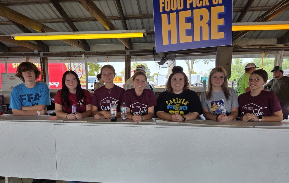 A photograph of seven high school students standing behind a white counter at what appears to be a food booth at a fair or outdoor event. Six of the seven students are wearing shirts related to FFA (Future Farmers of America) or a Carlyle school club. They are all smiling and posed behind the counter, with some holding drinks or small items. Above them, a large blue and yellow sign reads "FOOD PICK UP HERE." The structure has a metal roof and fluorescent lighting. People, fair rides, and vehicles are visible in the background.