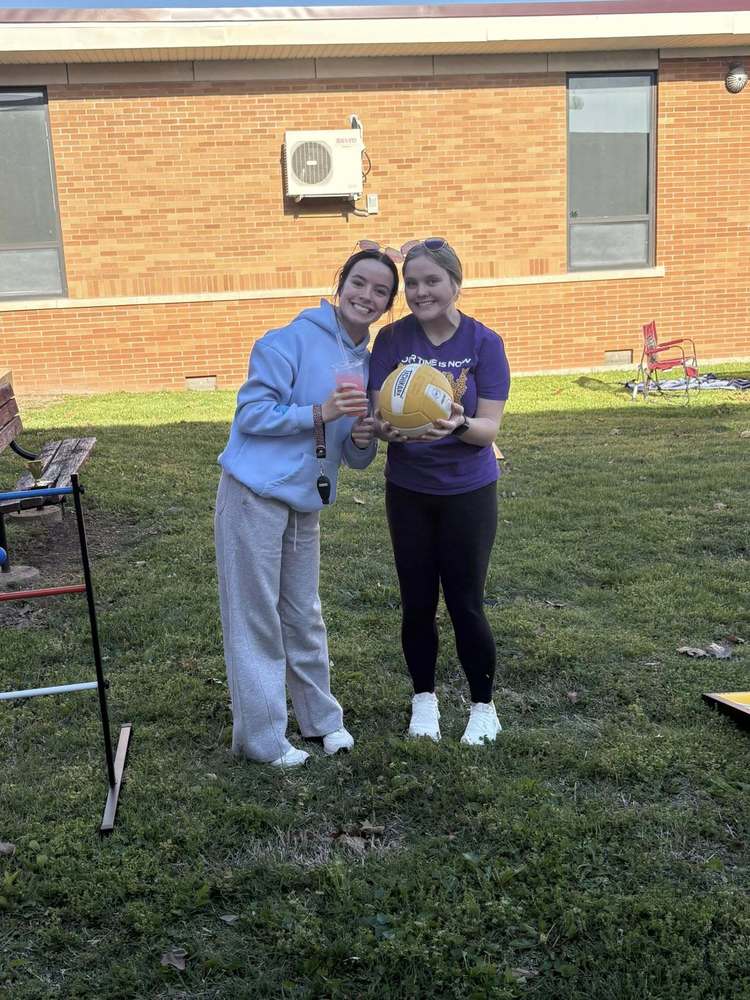 2 female students smile for a photo. One students holds a volleyball and the other a soda.