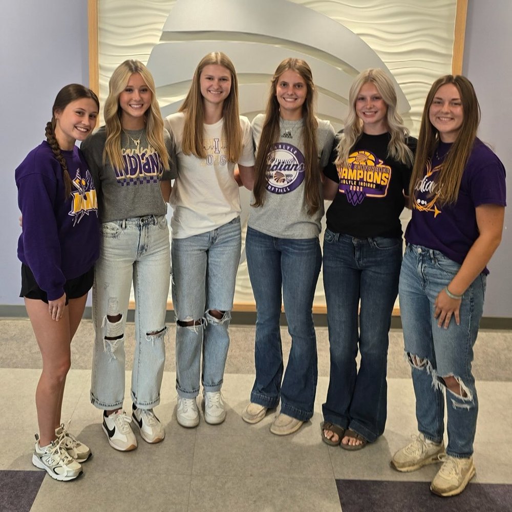 A photo of six high school girls standing together in a hallway.They are positioned in front of a wall with a modern, wave-like textured white and light purple art installation or paneling.