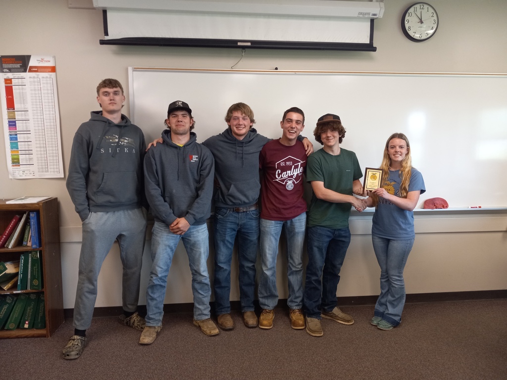 A photo of 5 team members smiling when receiving an award from a blonde girl. They are in a classroom setting.