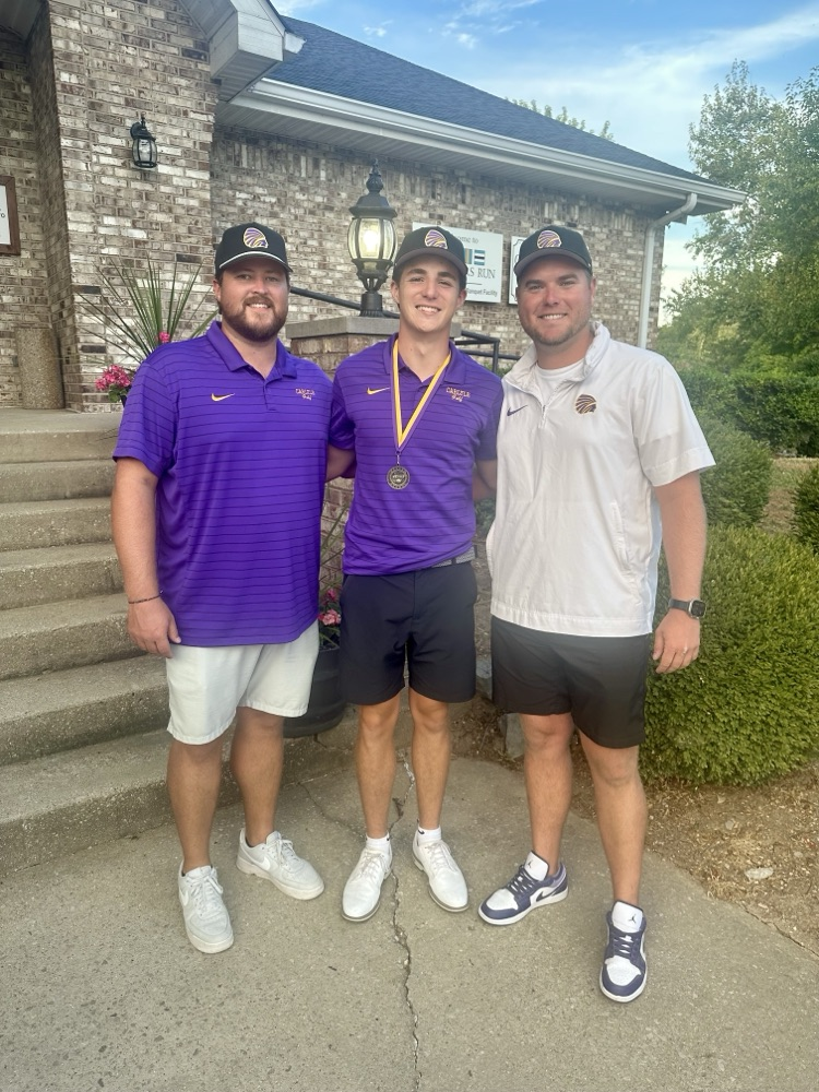 Two coaches posing with a golfer wearing a medal outside a brick building.