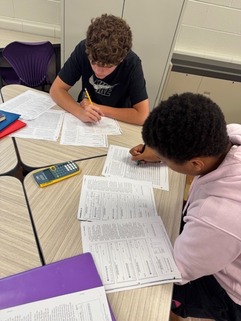 2 male students sit at a desk with worksheets and a calculator