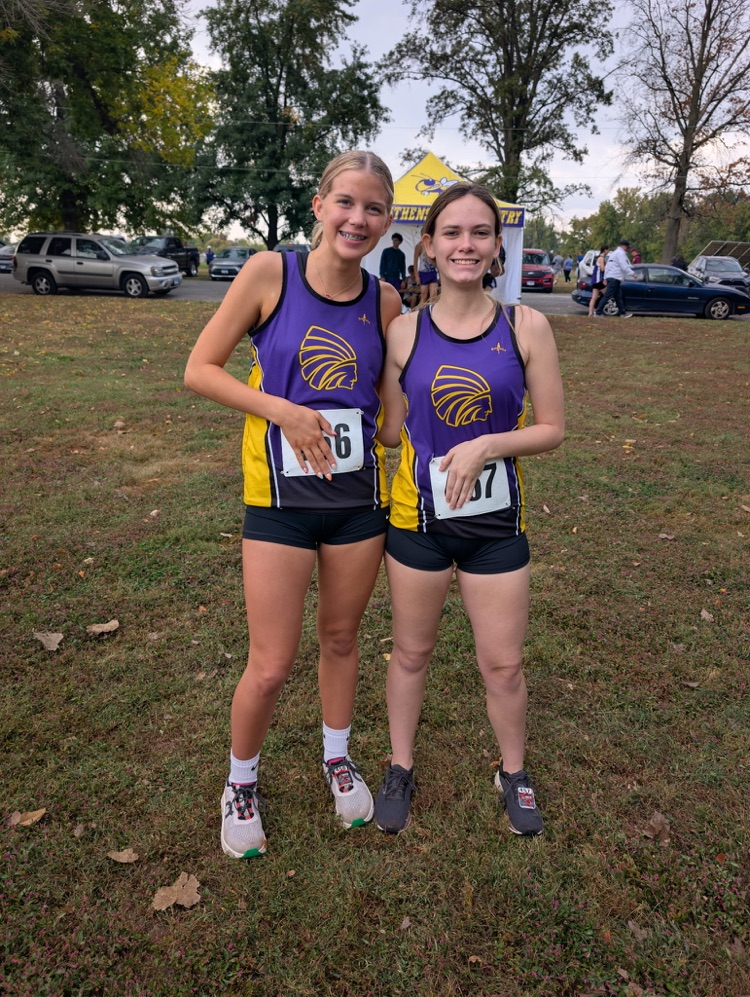 2 female cross country runners smile for a photo in an outdoor grassy area