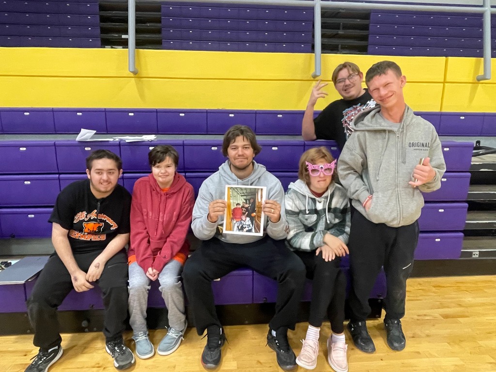 Life skills students and coach Taylor smile for a photo on the bleachers. Coach Taylor holds a picture of them doing their prank.