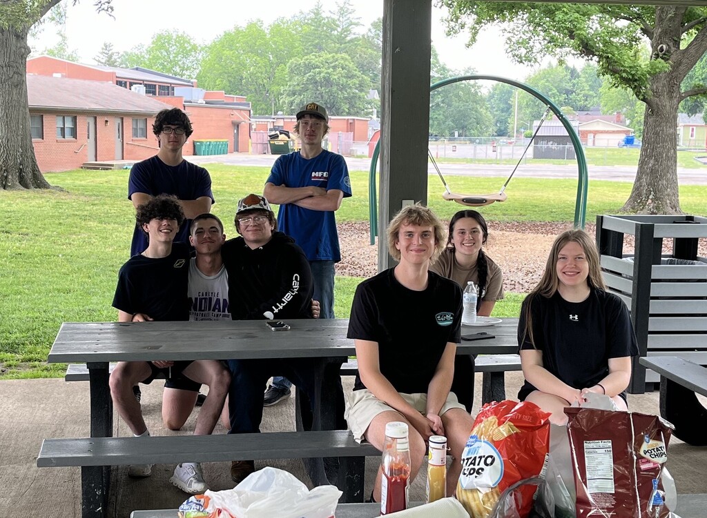 A group of eight teenagers posing together at a picnic table under a park pavilion. Bags of potato chips, paper towels, and condiments sit on the table in the foreground. In the background, there is a green grassy park area with trees, a playground with a swing set, and a brick building.