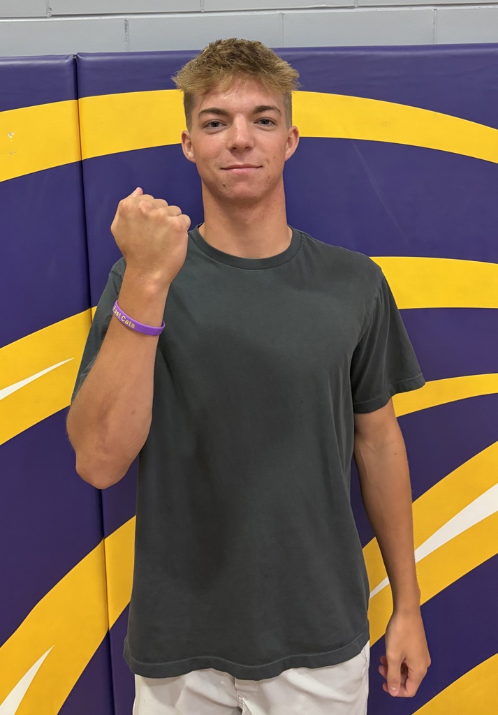 A young man stands in front of a purple and gold wrestling mat backdrop, wearing a dark gray T-shirt and white shorts. He smiles slightly while raising his left fist, showing a purple wristband that reads “Fast Cats.”