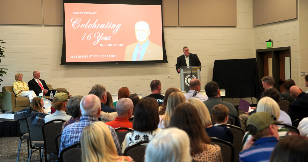 board chair kerry miller addresses a large crowd gathers to celebrated scott cowarts retirement. mr. and mrs. cowart are seated on stage