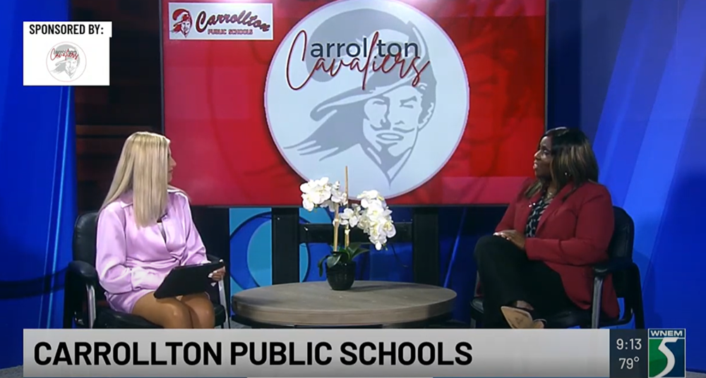  A television interview features a representative from Carrollton Public Schools seated across from a news anchor in a studio. The Carrollton Cavaliers logo is prominently displayed on a screen behind them, along with the text “Carrollton Public Schools.” A small potted plant is placed on the table between them. A sponsorship graphic in the top left corner shows the Carrollton Public Schools logo. The TV channel WNEM 5 logo and timestamp (9:13, 79°F) appear in the bottom right corner.