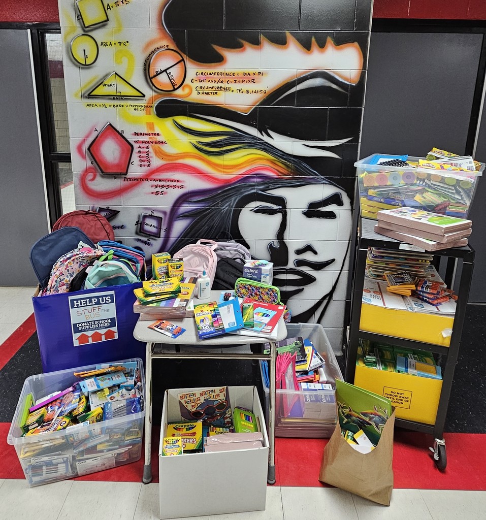 A vibrant display of donated school supplies collected during the “Stuff the Bus” event sits in front of a colorful mural featuring mathematical formulas and a stylized face wearing a feathered headdress. The supplies include backpacks, crayons, notebooks, folders, pencils, markers, and various classroom essentials neatly organized in bins, boxes, and on a desk and cart. A blue bin with a “Help Us Stuff the Bus” sign is prominently placed among the items.