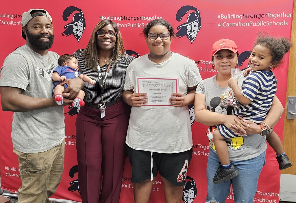 A smiling student holds a certificate of recognition while standing with family members and a school staff member in front of a red Carrollton Public Schools backdrop decorated with the Cavalier logo and the hashtag #BuildingStrongerTogether. One adult is holding a baby, another adult is holding a small child with candy, and everyone is smiling proudly for the photo.