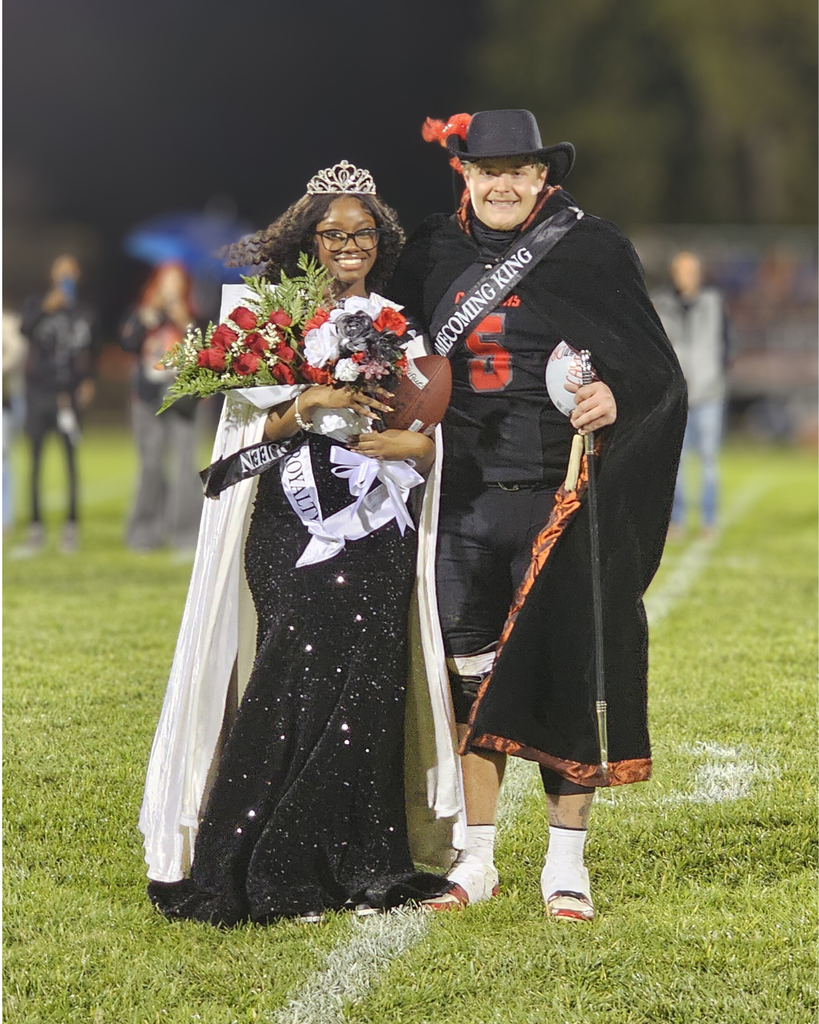 Two smiling students stand together on a football field at night, crowned as Homecoming King and Queen. The queen wears a sparkly black gown, a tiara, and a white cape, holding a large bouquet of red and white flowers. The king wears a football uniform with a black cape, crown, and cowboy-style hat, holding a cane and football. Both wear sashes reading “Homecoming King” and “Homecoming Queen,” with the stadium lights and blurred crowd in the background.