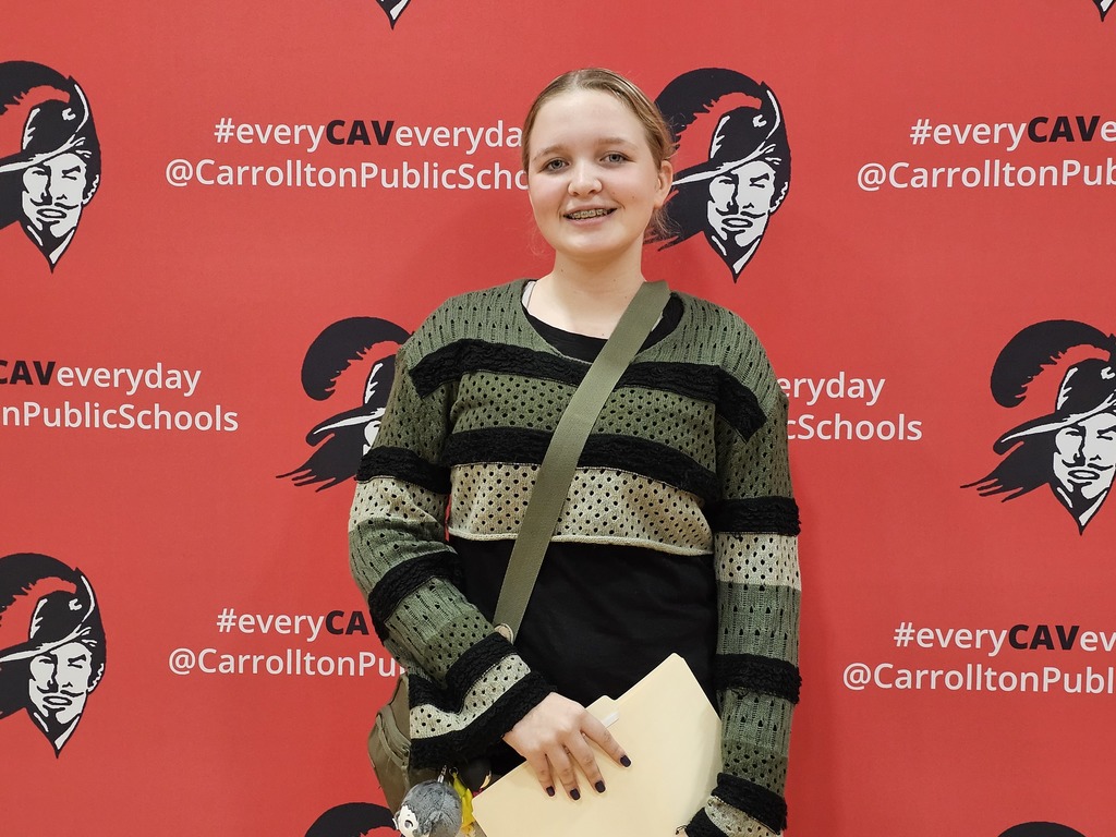 A smiling student stands in front of a bright red Carrollton Public Schools backdrop featuring the Cavalier logo and the hashtag #everyCAVeveryday. The student is wearing a green and black striped sweater over a black shirt, with a crossbody bag and holding a folder in one hand.