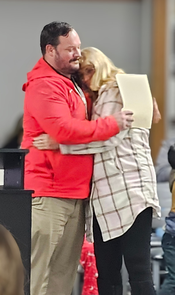 Two adults share a hug during a school recognition event. One person wears a red Cavaliers Athletics jacket, while the other holds a certificate and wears a light plaid jacket. They stand near a podium with a seated audience visible in the background, capturing a warm, appreciative moment.