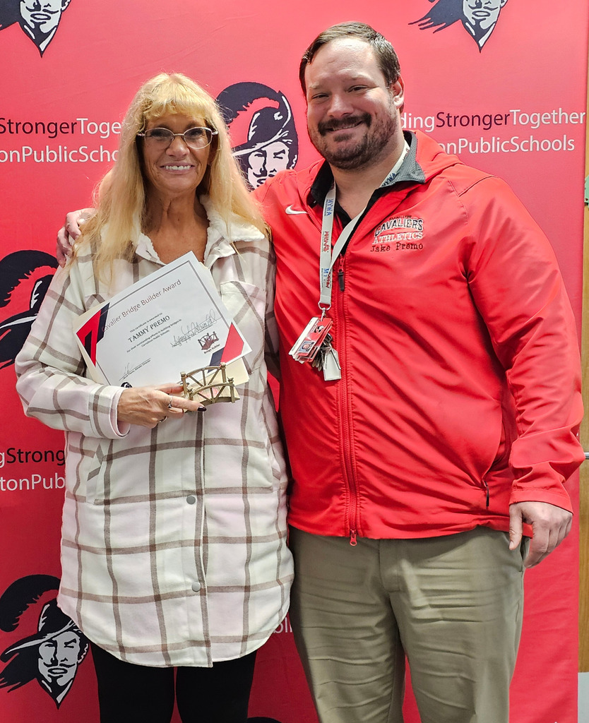 Two adults stand smiling in front of a red Carrollton Public Schools backdrop. One person holds a certificate and a small bridge-shaped award, while the other wears a red Cavaliers Athletics jacket and stands with an arm around them, suggesting an award presentation or recognition moment.