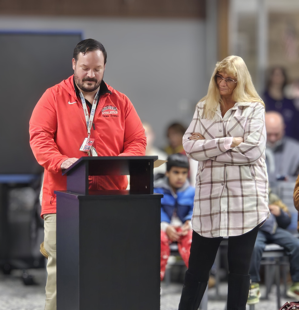 A staff member wearing a red Cavaliers Athletics jacket stands at a black podium reading from notes while another adult stands beside them with arms crossed, listening. They are indoors in front of a seated audience, with several students visible in the background, suggesting a school meeting or assembly.