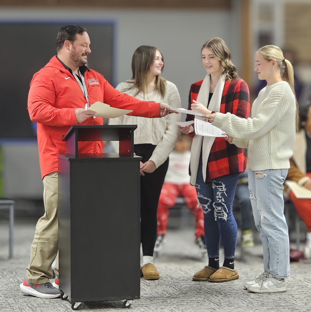 An adult wearing a red Cavaliers Athletics jacket stands at a podium and hands recognition certificates to three students during a school assembly. The students stand in a line smiling as they receive their certificates, with other seated students visible in the background.