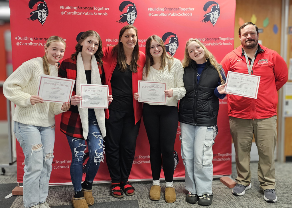 Five students and two adults stand smiling in front of a red Carrollton Public Schools “Building Stronger Together” backdrop. The students hold recognition certificates, while an adult in the center and another adult in a red Cavaliers Athletics jacket stand alongside them, capturing a celebratory student recognition moment.