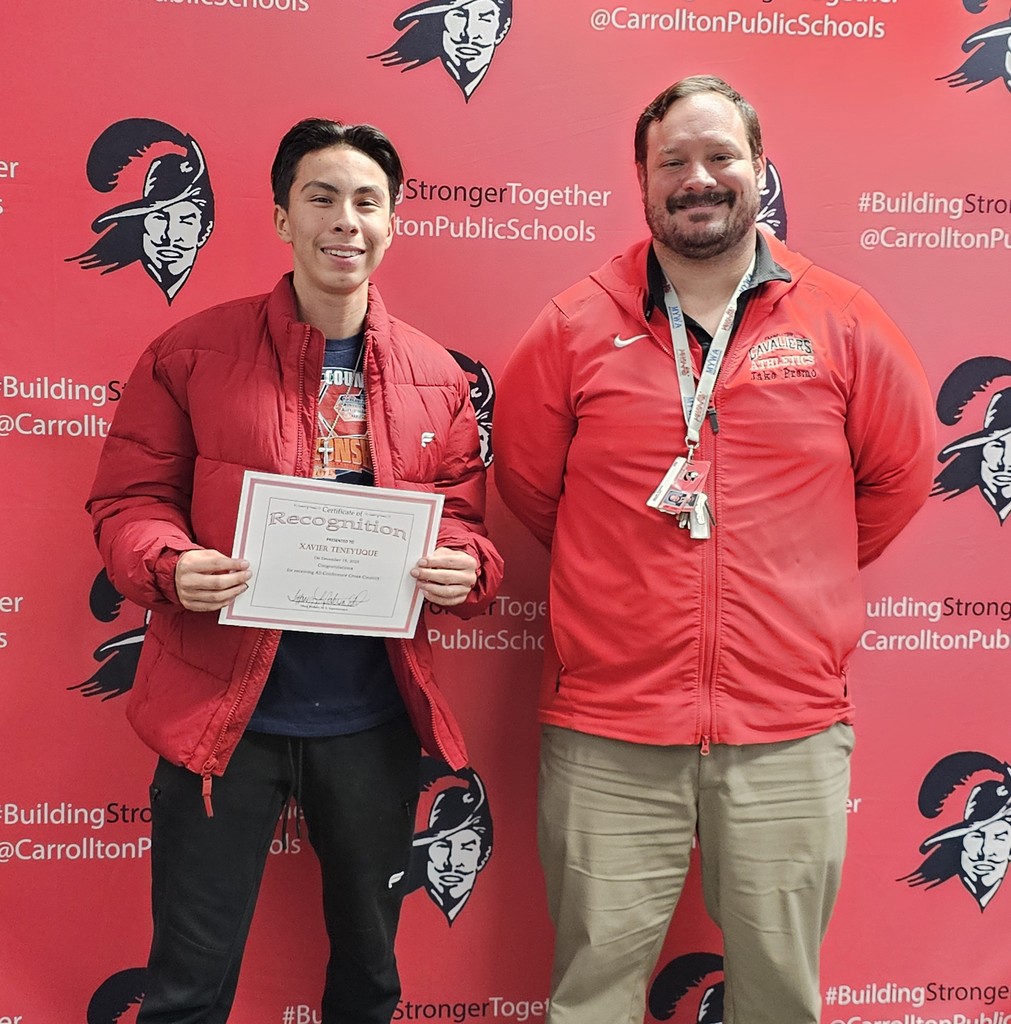 A high school student stands smiling and holding a recognition certificate next to an adult wearing a red Cavaliers Athletics jacket. They pose in front of a red Carrollton Public Schools backdrop with Cavalier logos and “Building Stronger Together” text, capturing a student-athlete recognition moment.