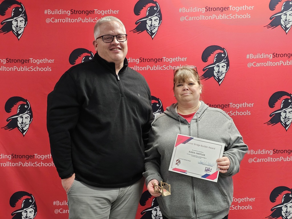 Two Carrollton Public Schools staff members stand in front of a red CPS backdrop featuring the Cavalier logo and the hashtag #BuildingStrongerTogether. On the left, a man wearing glasses and a black pullover smiles at the camera. On the right, a woman holds a Cavalier Bridge Builder Award certificate and a small bridge-shaped award.