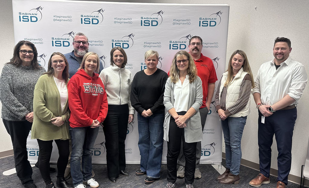 Ten adults stand together in front of a step-and-repeat backdrop displaying the Saginaw ISD logo and the text “#SaginawISD @SaginawISD.” The group is arranged in a single row, smiling at the camera in a conference or meeting room setting. They are dressed in casual-to-business-casual clothing including sweaters, jeans, a red Omni High School hoodie, a red polo shirt, and a white button-down shirt. The neutral-colored wall and carpeted floor suggest an indoor professional event or training session.