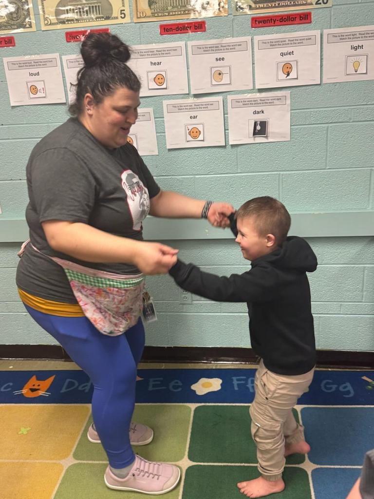 A teacher and a young child hold hands and smile as they dance together on a colorful classroom rug. The teacher wears a gray T-shirt and patterned apron, while the child is dressed in a black hoodie and tan pants, standing barefoot. Behind them, a classroom wall displays labeled pictures and words