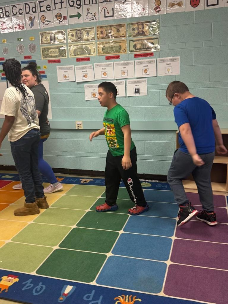Three students and a teacher move in a line across a colorful classroom rug during an activity. The students follow one another while stepping or walking, and the teacher stands nearby guiding them. The wall behind them displays alphabet cards, labeled pictures with words