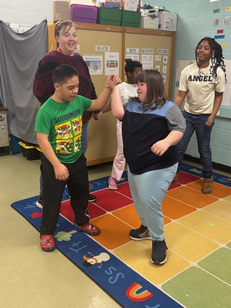 A teacher and several students stand on a colorful classroom rug participating in a movement activity. Two students hold hands and spin or dance together in the center, smiling and engaged, while other students and the teacher watch and encourage them. Behind them are classroom cabinets and posters with learning materials, creating a supportive and interactive learning environment.