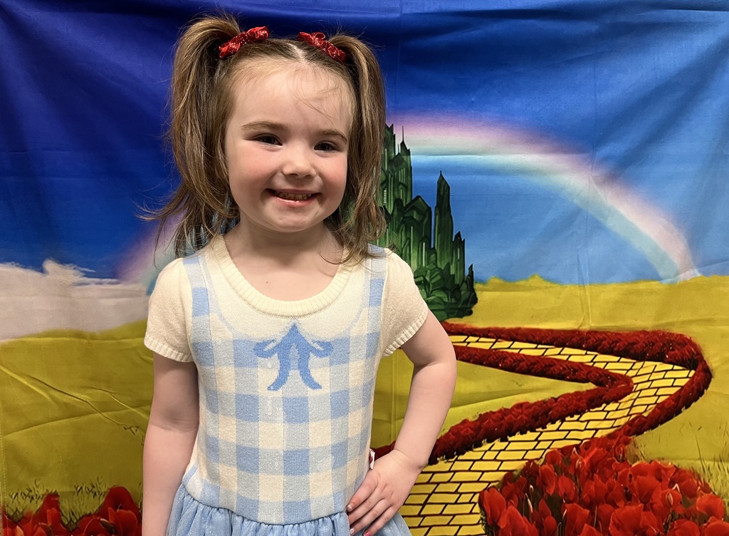 A smiling young child with brown hair in pigtails tied with red bows stands in front of a colorful Wizard of Oz–themed backdrop. She is wearing a light blue and white gingham dress with a white shirt underneath, resembling Dorothy’s outfit. Behind her is a painted scene featuring a bright yellow brick road, red flowers, a rainbow, and the green Emerald City in the distance.
