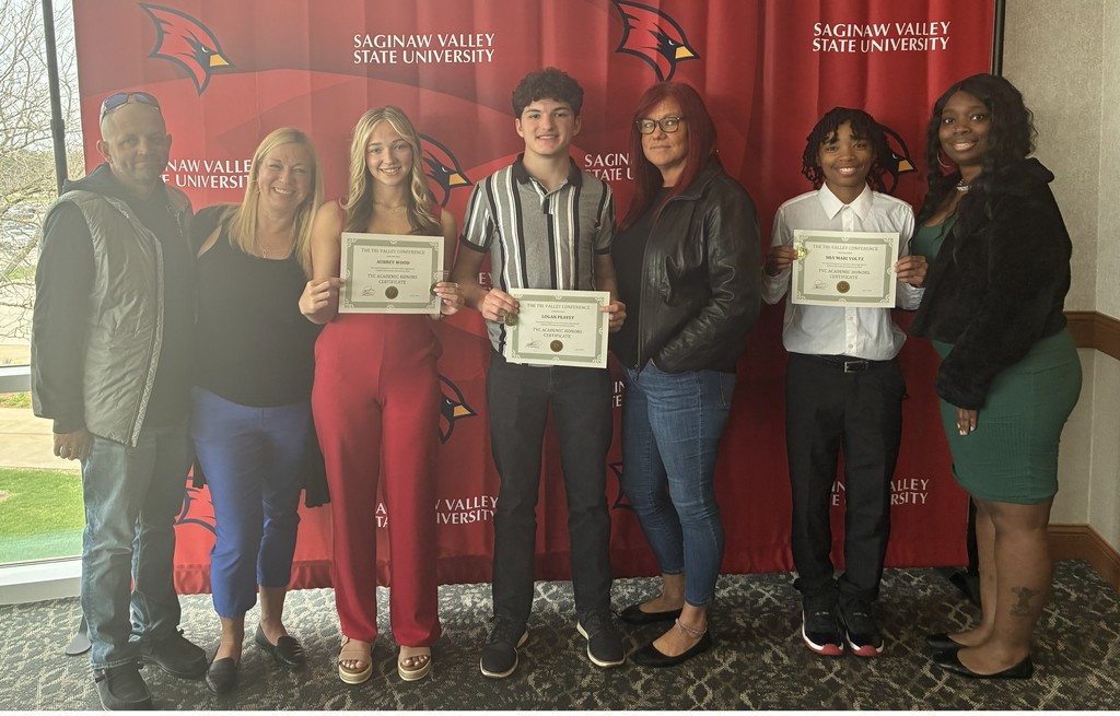 Seven people stand together in front of a red Saginaw Valley State University backdrop with Cardinal logos. In the center, three high school students smile and hold “TVC Academic Honors Certificate” awards. They are surrounded by adults—likely family members or staff—standing on both sides, also smiling. The group is dressed in a mix of casual and semi-formal attire, posing proudly to celebrate the students’ academic achievements.