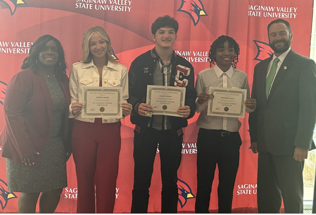 Five people stand in front of a red Saginaw Valley State University backdrop with Cardinal logos. Three high school students in the center smile while holding certificates labeled “TVC Academic Honors Certificate.” The students are dressed in semi-formal attire. An adult woman stands on the far left and an adult man in a suit stands on the far right, both smiling as they pose with the students to celebrate their academic achievement.