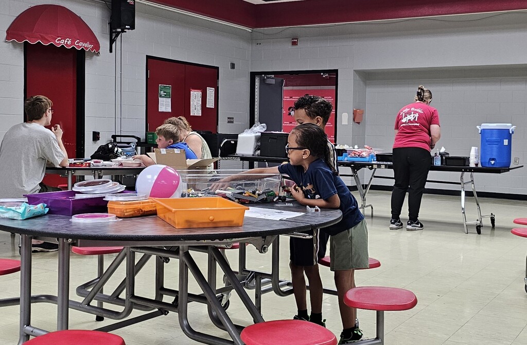 Two children explore hands-on activities at a cafeteria table during a community event, reaching into a bin filled with building materials and toys. Other children, families, and volunteers participate in activities and visit resource tables in the background.