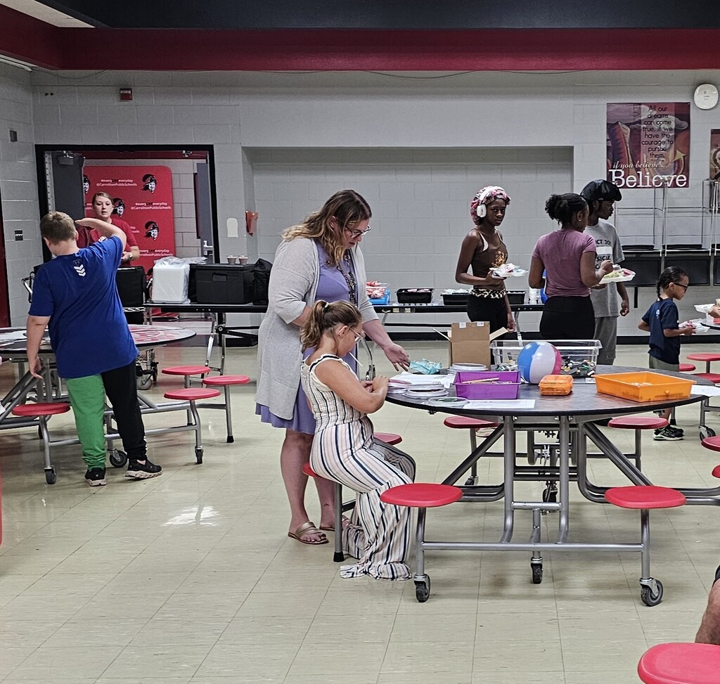 A child works on a hands-on activity at a cafeteria table while an adult stands nearby offering support. Other children and volunteers gather around tables in the background, enjoying activities and refreshments during a community event.