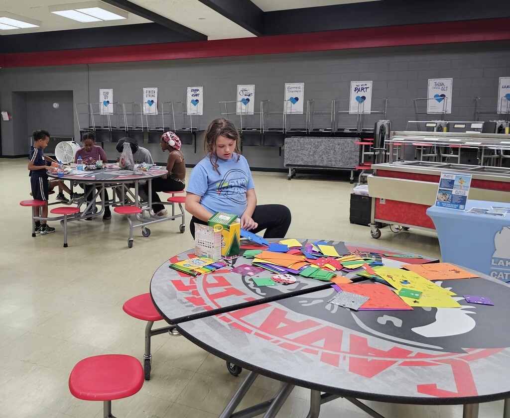 A child works on a colorful craft project at a cafeteria table covered with paper, markers, crayons, and other art supplies during a community event. Other children create projects together at a nearby table in the background.