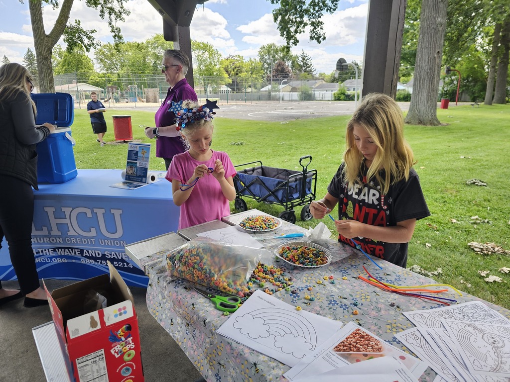Two children make colorful cereal-bead bracelets using pipe cleaners at an outdoor activity table. Bowls and bags of cereal, craft supplies, and rainbow activity sheets cover the table, with Lake Huron Credit Union representatives and a resource table nearby.