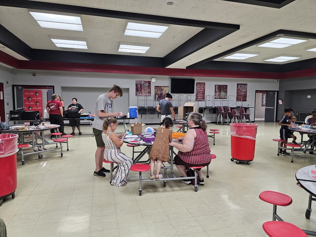 Children, families, and volunteers gather around cafeteria tables for hands-on activities during a community event. In the center, children work on crafts with adults while other participants visit activity and resource tables throughout the room.