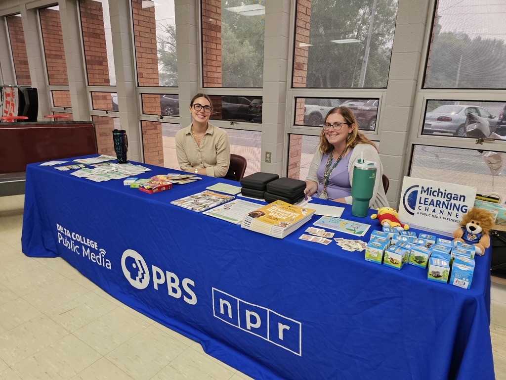 Two smiling representatives sit behind a blue PBS and NPR table at a community event. The table displays Michigan Learning Channel educational resources, books, activity materials, giveaways, and small stuffed animals. A Michigan Learning Channel sign is displayed on the right side of the table.