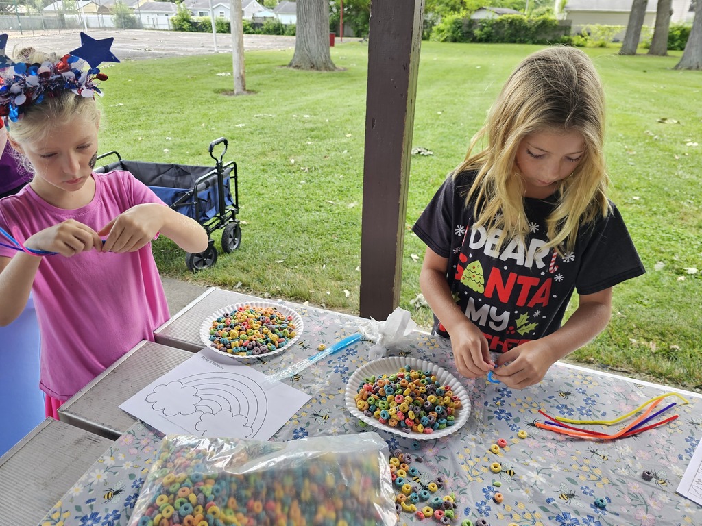Two children make colorful cereal-bead bracelets using pipe cleaners at an outdoor activity table. Bowls of brightly colored cereal, craft supplies, and a rainbow activity sheet are spread across the table.