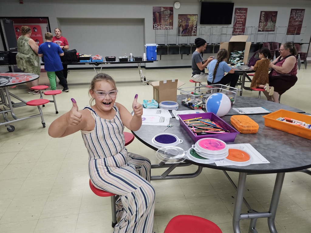 A smiling child gives two thumbs up, showing purple ink on her thumbs while seated at a craft table during a community event. Colorful ink pads, markers, activity sheets, and other craft supplies cover the table as families and volunteers participate in activities in the background.