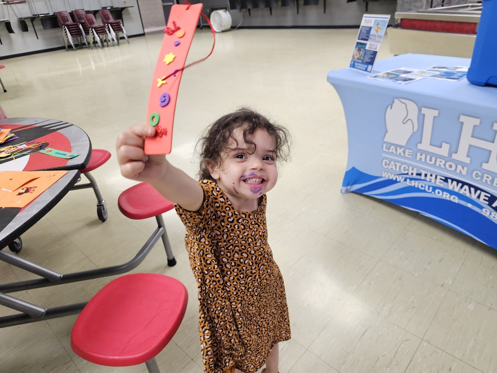 A smiling young child proudly holds up a red handmade bookmark decorated with colorful beads and string during a community event. Craft supplies are visible on a nearby table, with a Lake Huron Credit Union resource table in the background.