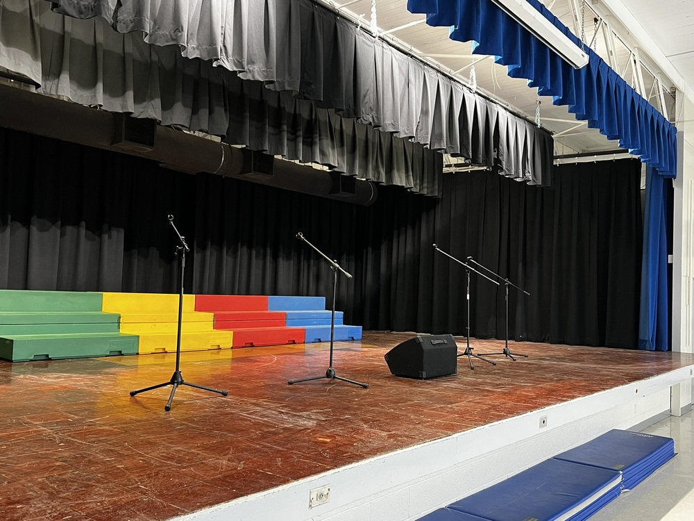 A photograph of the MCES performing arts stage with colored risers on the stage and 4 microphone stands.