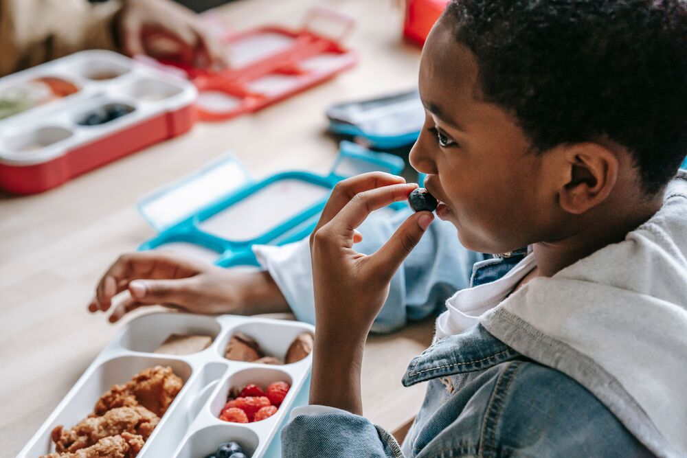 African American male student in a denim jacket eating lunch in the cafeteria (tray appears to have chicken tenders, raspberries, and blueberries on it)
