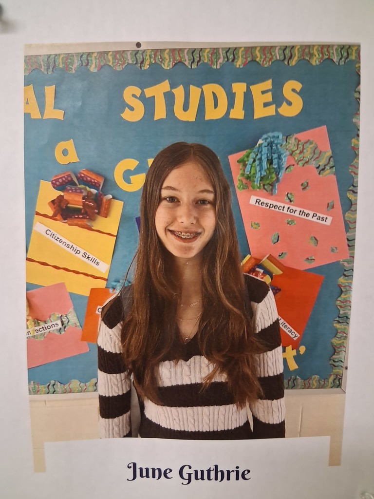 June Guthrie, wearing a black and white striped shirt, stands in front of a colorful bulletin board. 