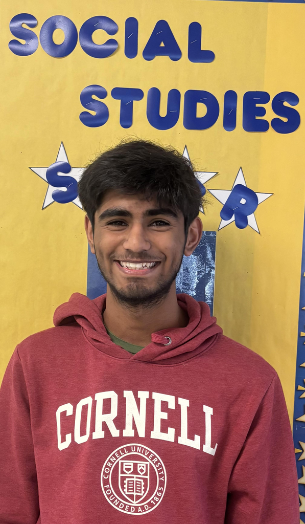 Ayush is wearing a Cornell hoodie and stands in front of a bulletin board with the words Social Studies Star in blue letters with a yellow background.