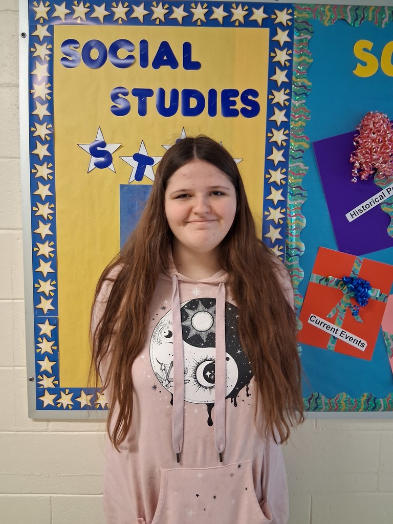 Ava is smiling and stands in front of a yellow bulletin board with "Social Studies Star" in blue letters stapled to the board. There is a blue border with yellow stars framing the board.