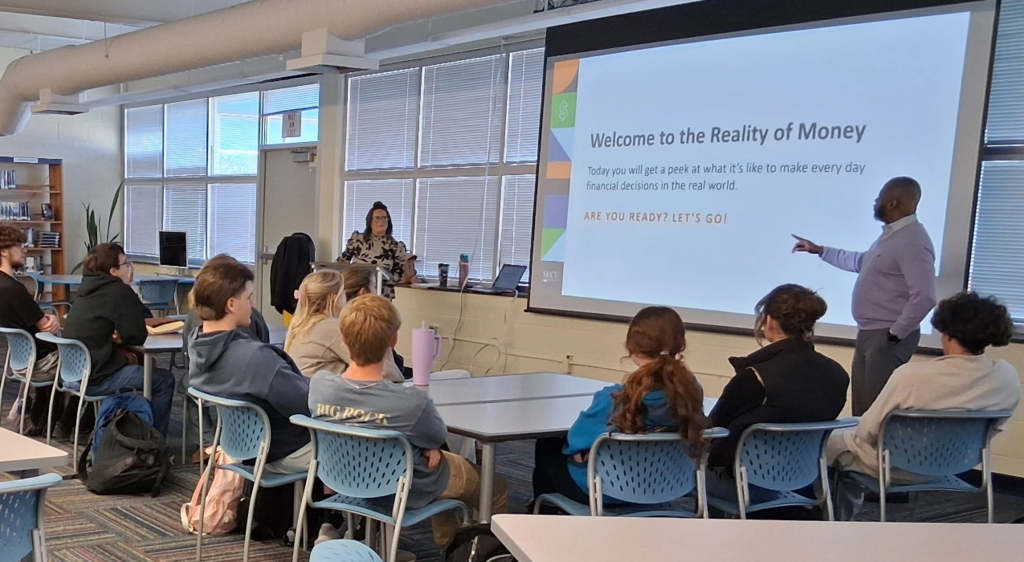 Presenters Brandy Pittman and Jason Wilson stand in front of students. They share a slide that states "Welcome to the Reality of Money."