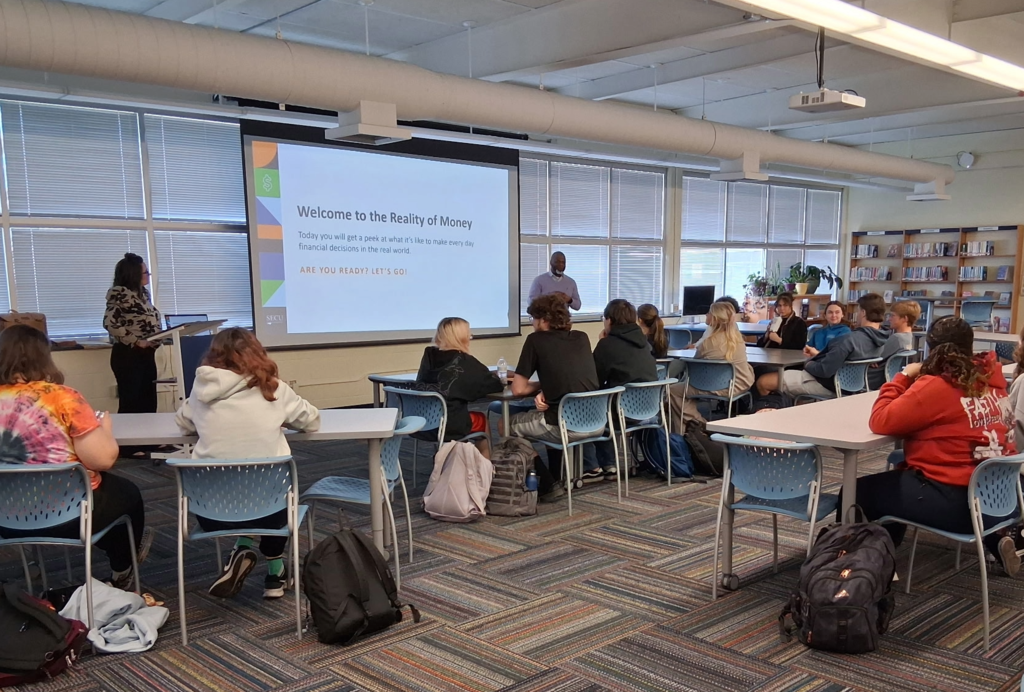 Presenters Brandy Pittman and Jason Wilson stand in front of students. They share a slide that states "Welcome to the Reality of Money."