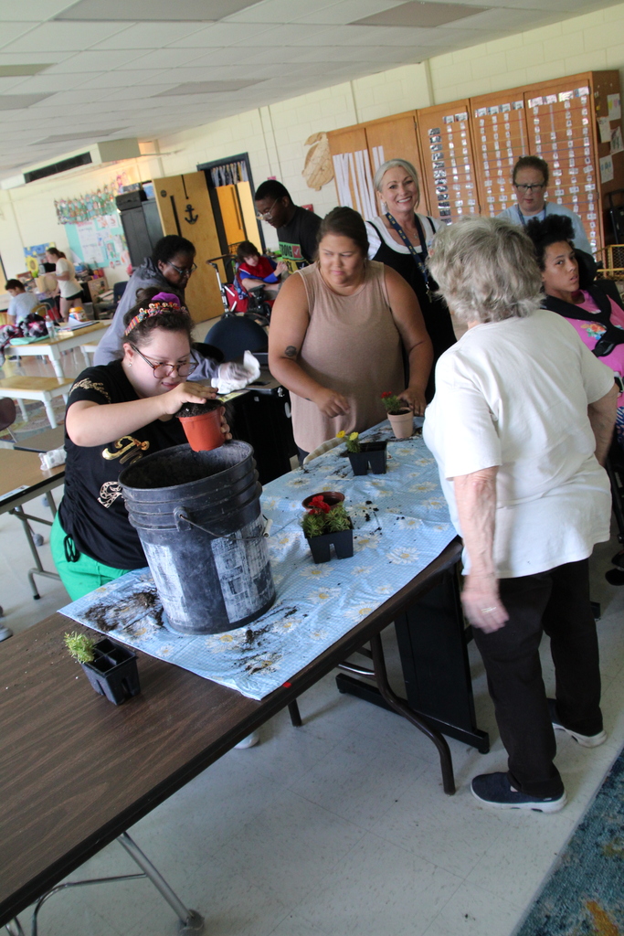 The class plants flowers on long tables. Buckets of dirt and protective covers are on the tables along with flowers and pots.