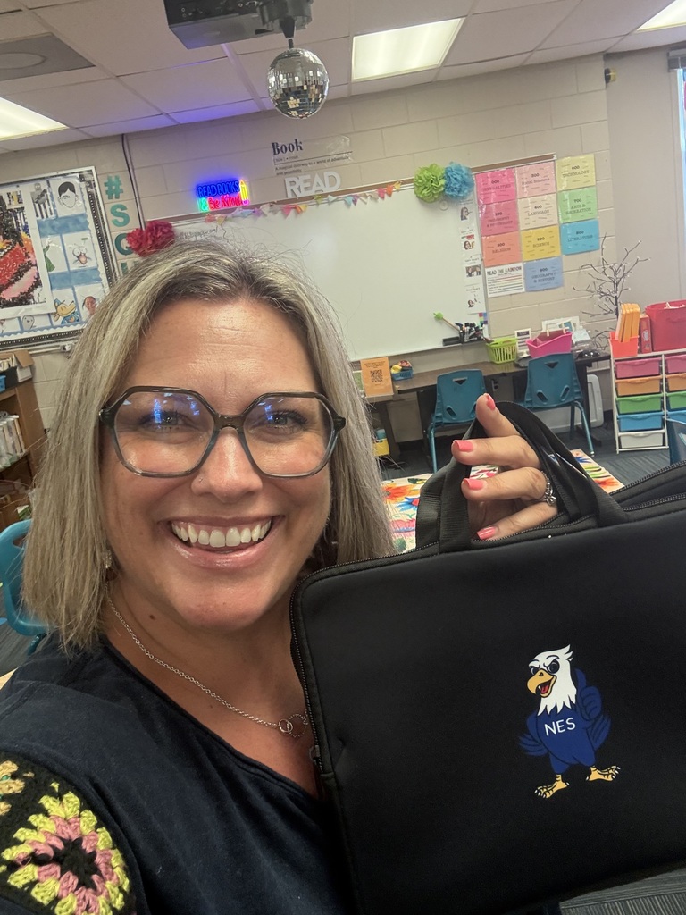 Mrs. Golightly in the library holding a Chromebook carrying bag with the NES hawk logo on it