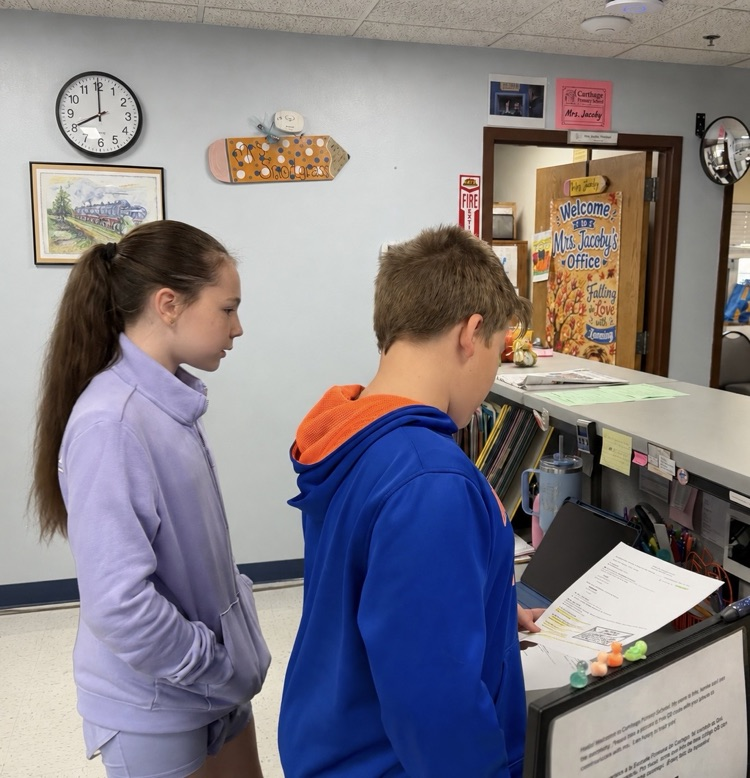 two students standing in front of secretary desk reading announcements